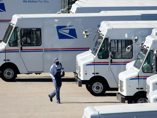 USPS postal worker, in a blue uniform and cap, walks past a line of parked white postal trucks with red and blue stripes, conveying a busy workday