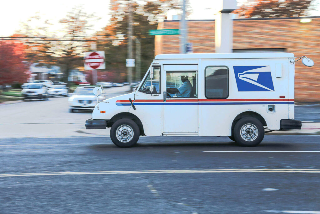 White USPS mail delivery truck with postal worker driving through residential street, symbolizing the mail delivery service that postages.co stamps support and enable.